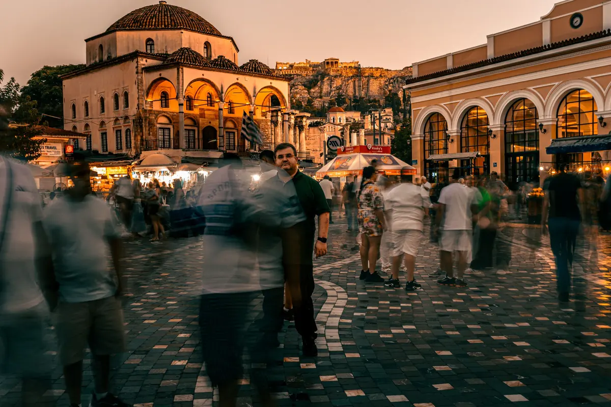 A busy Athens square at dusk, with an arcaded building and a domed historic building lit warm and people crossing the paving.
