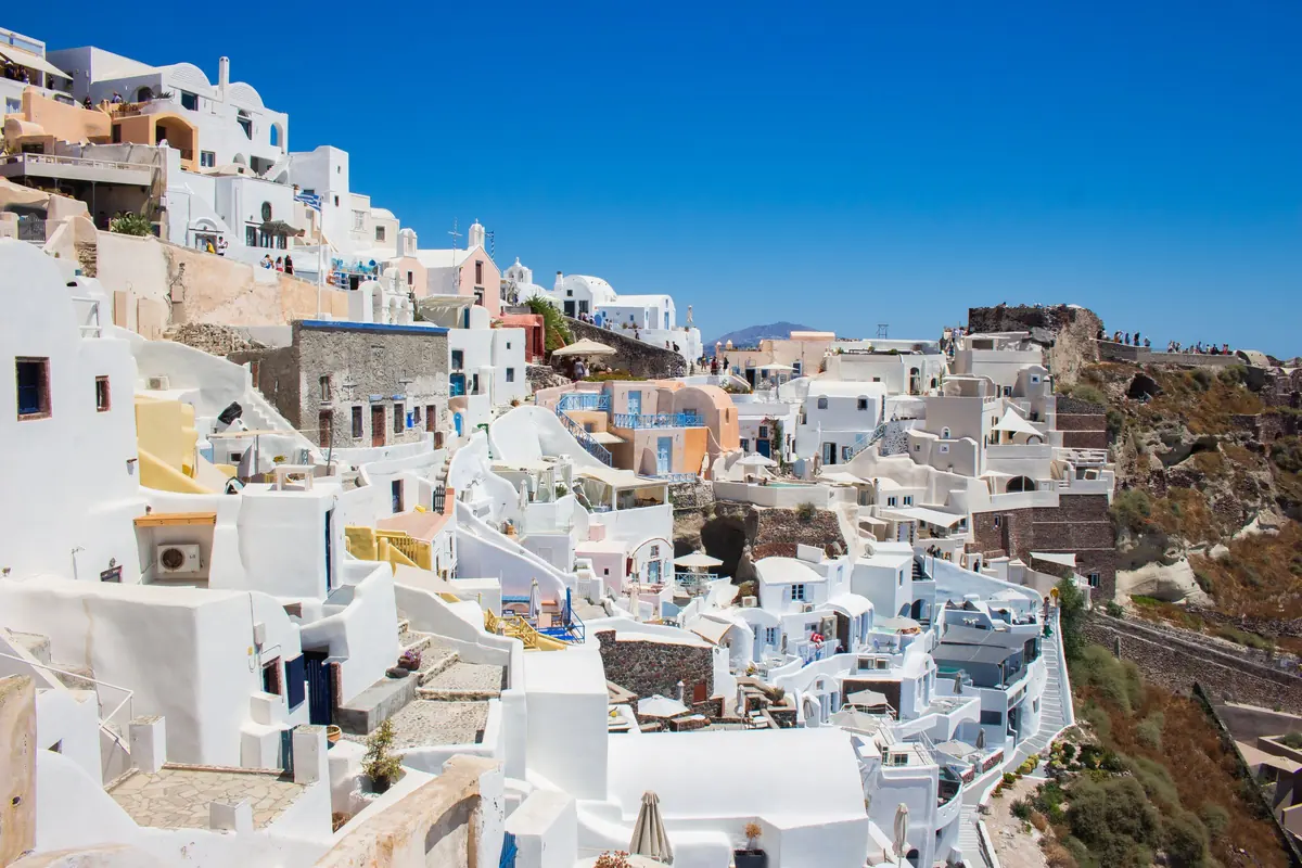 Whitewashed houses stepping down a steep slope above the sea on Santorini under a bright blue sky.