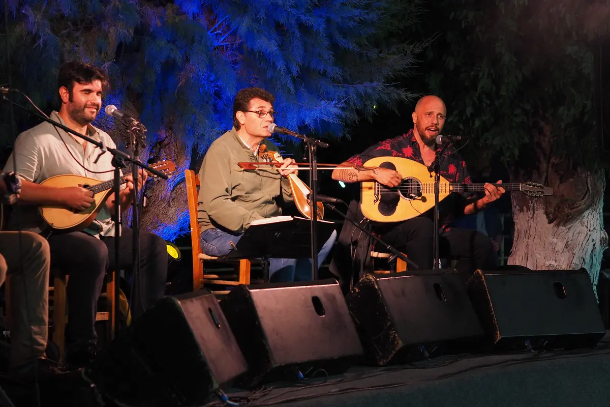 Three folk musicians seated on a stage lit blue, each with a stringed instrument and a microphone.
