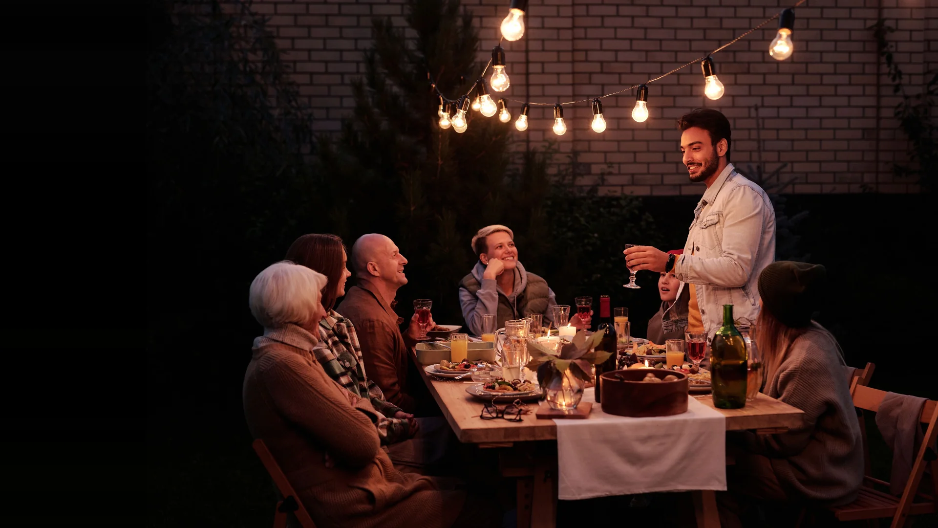 Representative photograph: a man stands with a raised glass at a long wooden table under strung lights at night, talking to guests who sit with candles, wine and plates in front of them.